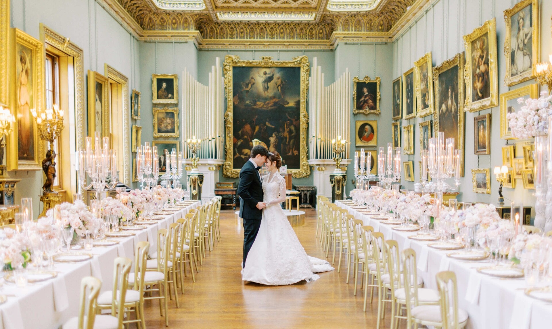 A couple in wedding attire shares a kiss in a grand, art-filled hall decorated for their reception by a renowned London wedding florist. The bride wears a white gown, and the groom is in a black suit. The space is in Somerley House and features two long banquet tables adorned with wedding flowers and numerous candles, with ornate paintings on the walls.